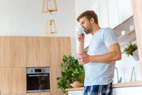 Selective focus of man drinking coffee and using smartphone at kitchen — Stock Photo