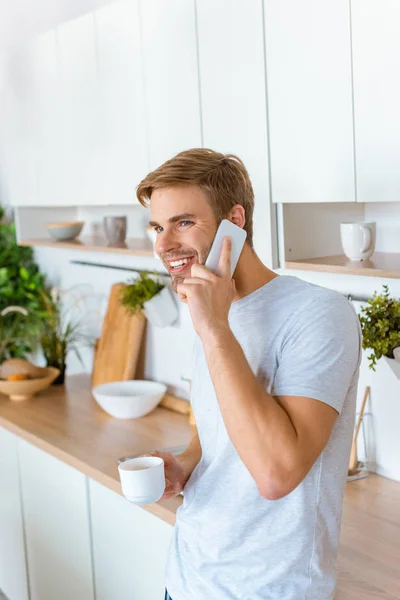 Усміхнений молодий чоловік з кавою розмовляє на смартфоні в ранковий час на кухні — стокове фото