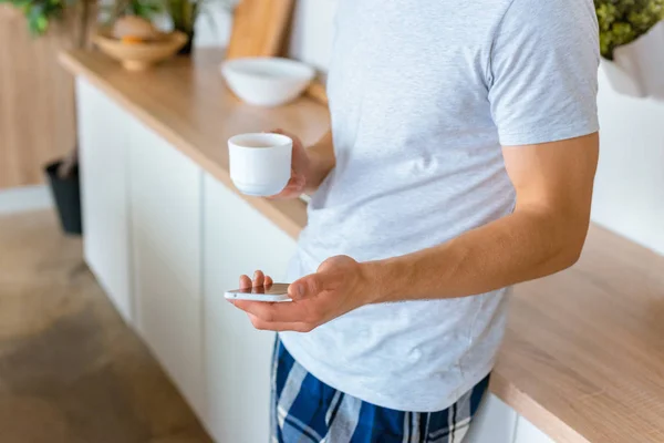 Imagen recortada de hombre joven con taza de café utilizando el teléfono inteligente en la cocina - foto de stock