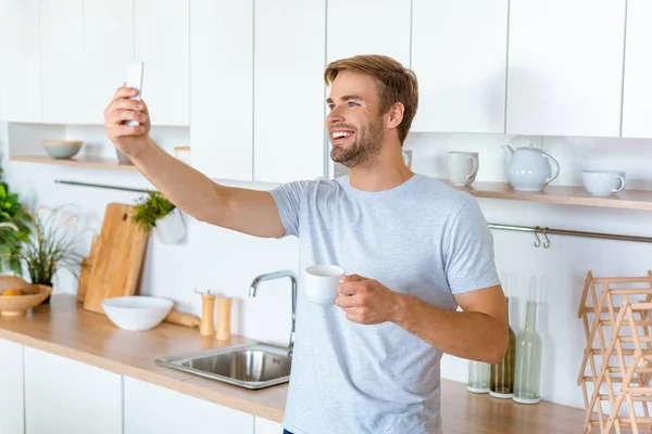 Красивый улыбающийся мужчина с кофейной чашкой делает селфи на смартфоне на кухне — стоковое фото