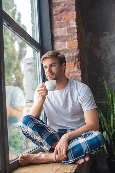 Enfoque selectivo de hombre joven sentado con taza de café en el alféizar de la ventana - foto de stock