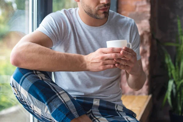 Imagen recortada de hombre joven sentado con taza de café en el alféizar de la ventana — Stock Photo