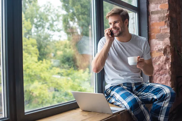 Heureux jeune homme avec tasse de café parler sur smartphone tout en étant assis sur le rebord de la fenêtre avec ordinateur portable à la maison — Photo de stock