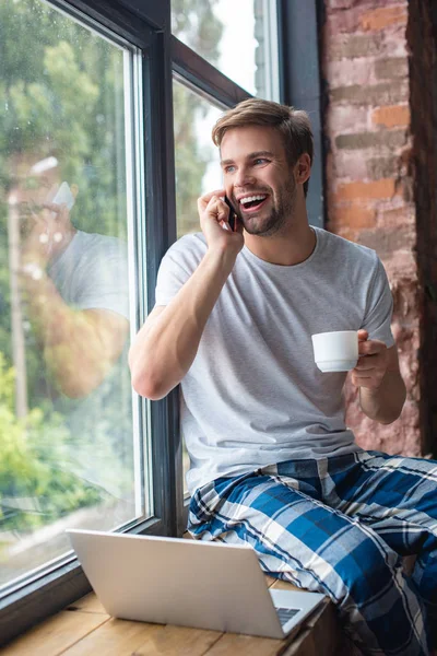 Jeune homme souriant avec tasse de café parlant sur smartphone tout en étant assis sur le rebord de la fenêtre à la maison — Photo de stock