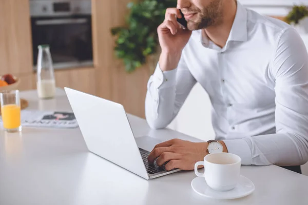 Imagen recortada de hombre de negocios hablando en el teléfono inteligente y el uso de ordenador portátil en la mesa de la cocina con taza de café - foto de stock
