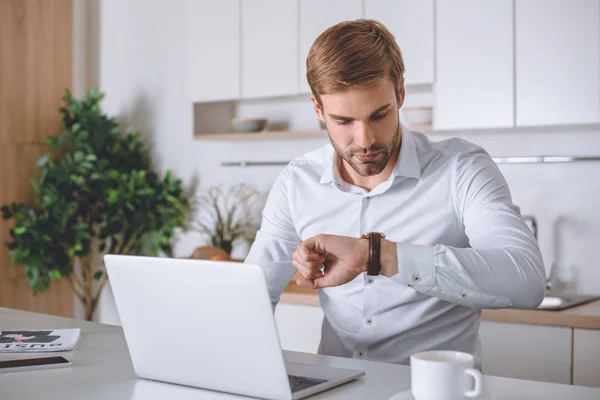 Jeune homme d'affaires regardant montre-bracelet à la table de cuisine avec ordinateur portable et café — Photo de stock