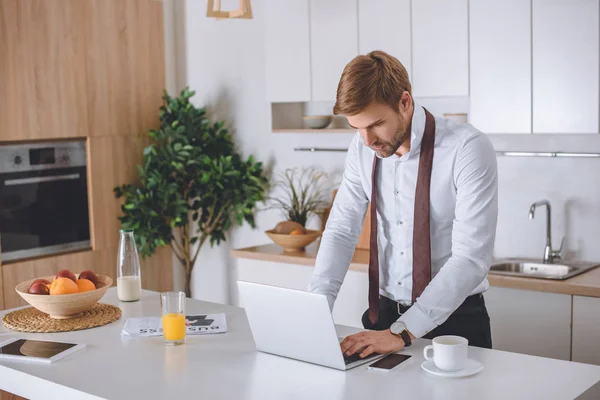 Joven hombre de negocios utilizando el ordenador portátil en la mesa de la cocina con teléfono inteligente y café - foto de stock