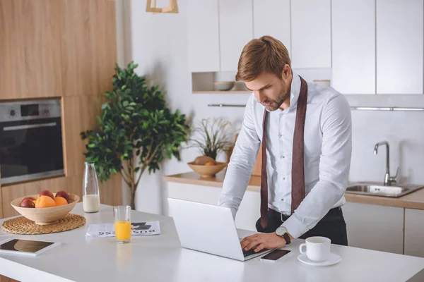 Jovem empresário usando laptop na mesa da cozinha com smartphone e café — Fotografia de Stock