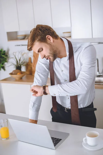 Seriöser Geschäftsmann, der am Smartphone telefoniert und auf eine Armbanduhr am Küchentisch mit Laptop und Kaffeetasse blickt — Stockfoto