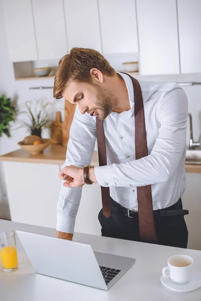 Homme d'affaires sérieux parlant sur smartphone et regardant montre-bracelet près de la table de cuisine avec ordinateur portable et tasse à café — Photo de stock