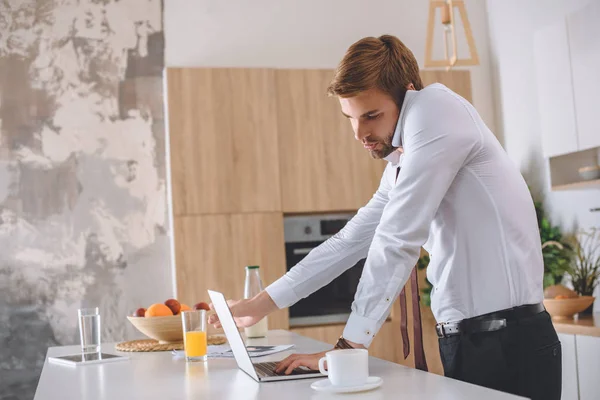 Confiante jovem empresário falando no smartphone e usando laptop na mesa da cozinha com xícara de café — Fotografia de Stock