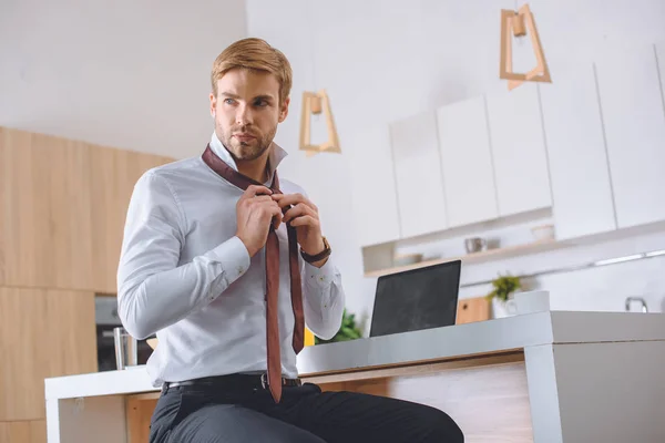 Confident young businessman tying necktie at kitchen table with laptop — Stock Photo