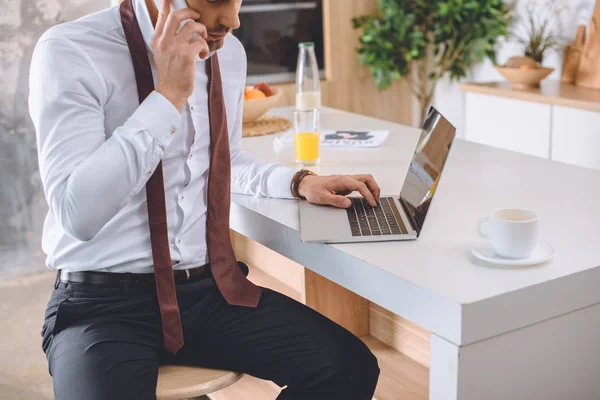 Cropped image of businessman in white shirt with necktie over neck talking on smartphone and working on laptop at kitchen — Stock Photo