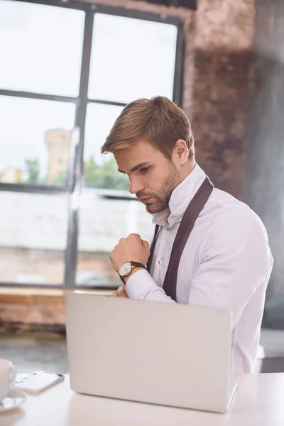Joven hombre de negocios guapo poniéndose un reloj de pulsera cerca de la computadora portátil - foto de stock