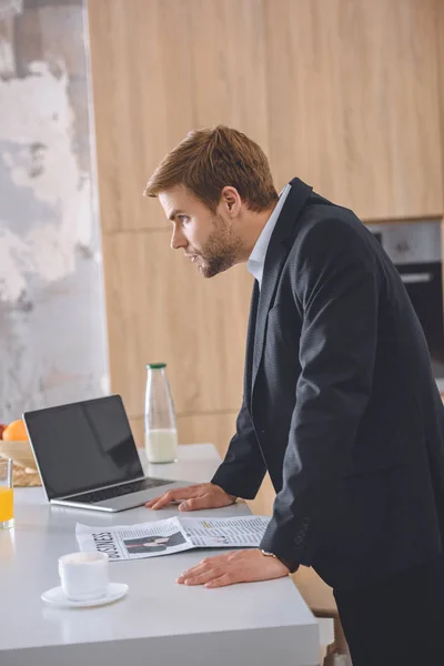 Serious businessman standing at kitchen table with laptop and business newspaper — Stock Photo