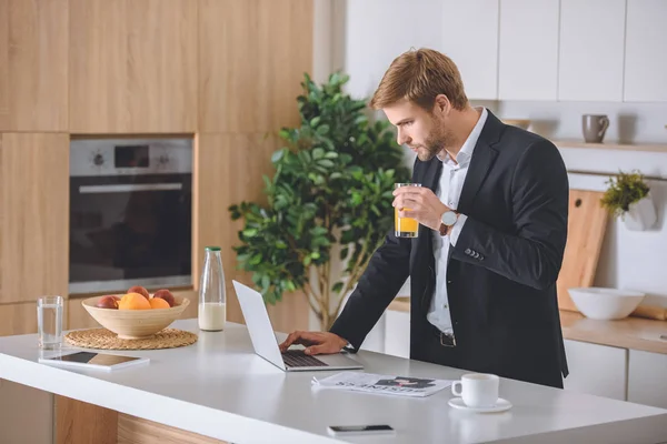 Enfocado hombre de negocios beber jugo fresco usando el ordenador portátil en la mesa de la cocina durante el desayuno - foto de stock