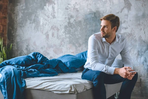 Молодий бізнесмен зі смартфоном в руці сидить на ліжку вдома — стокове фото