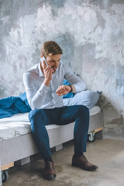 Jungunternehmer spricht auf Smartphone und schaut zu Hause im Schlafzimmer auf Armbanduhr — Stockfoto