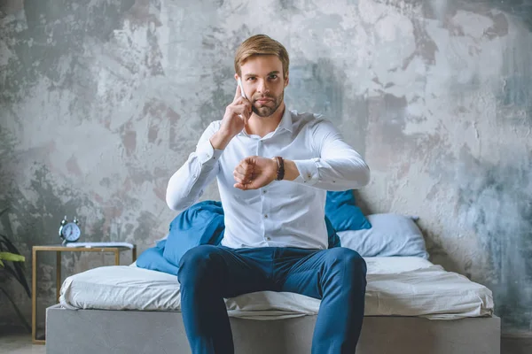 Homme d'affaires souriant parlant sur smartphone et regardant montre-bracelet dans la chambre à coucher à la maison — Photo de stock