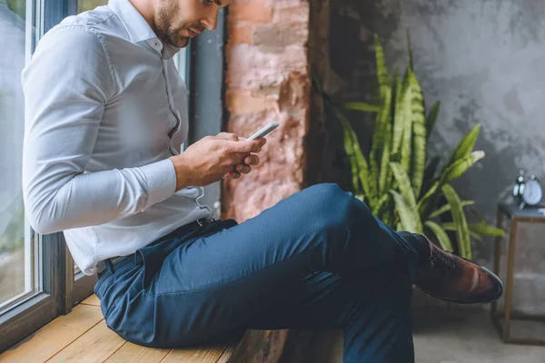 Immagine ritagliata di uomo d'affari utilizzando smartphone sul davanzale della finestra a casa — Foto stock