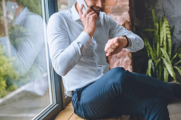 Imagen recortada de hombre de negocios hablando en el teléfono inteligente y mirando reloj de pulsera en casa - foto de stock