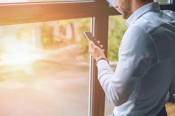 Imagen recortada de joven empresario usando teléfono inteligente cerca de ventanas con luz solar - foto de stock