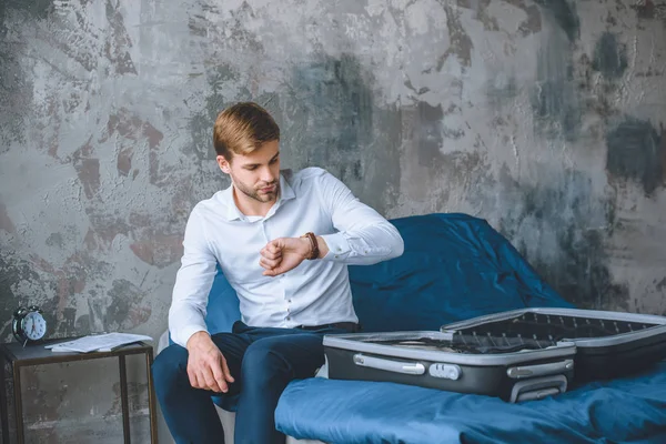 Junger Geschäftsmann schaut auf Armbanduhr, während er im Bett sitzt — Stockfoto