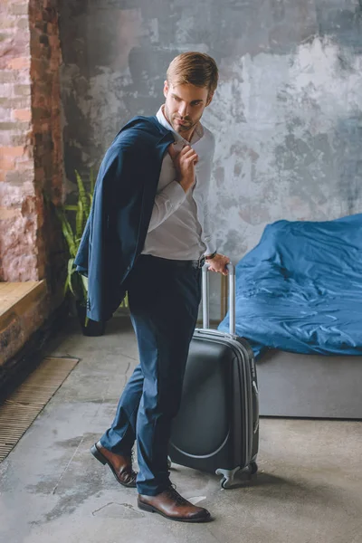 Молодий бізнесмен з піджаком над плечем, що несе валізу в спальні вдома — стокове фото