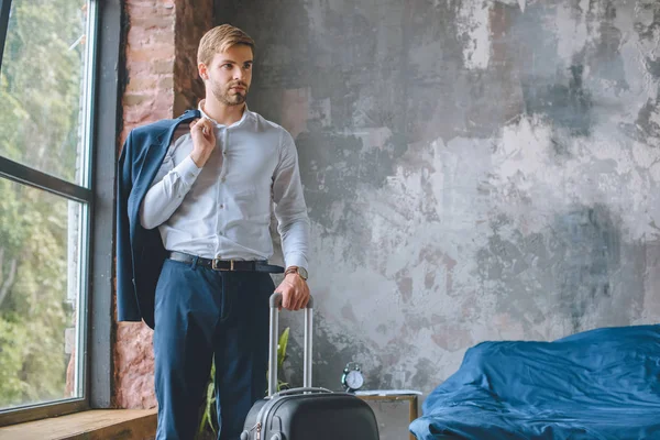 Впевнений чоловік мандрівник з курткою над плечем, що стоїть з валізою в спальні вдома — стокове фото
