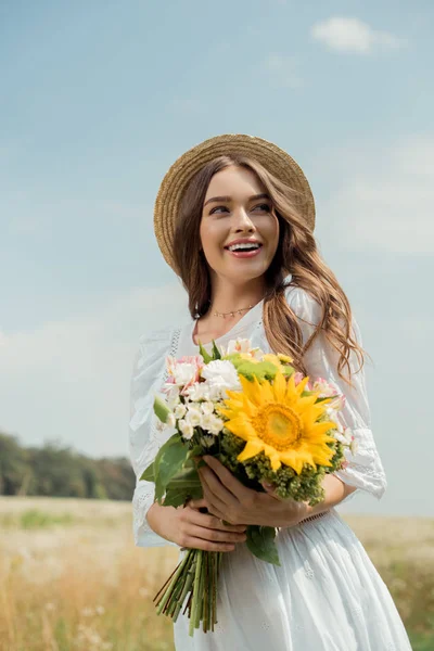 Portrait of cheerful woman in white dress with bouquet of wild flowers in field — Stock Photo