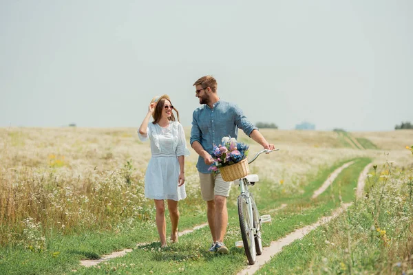 Smiling couple in sunglasses with retro bicycle in summer field with wild flowers — Stock Photo