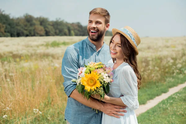 Retrato de hombre sonriente abrazando novia con ramo de flores silvestres en el campo de verano - foto de stock