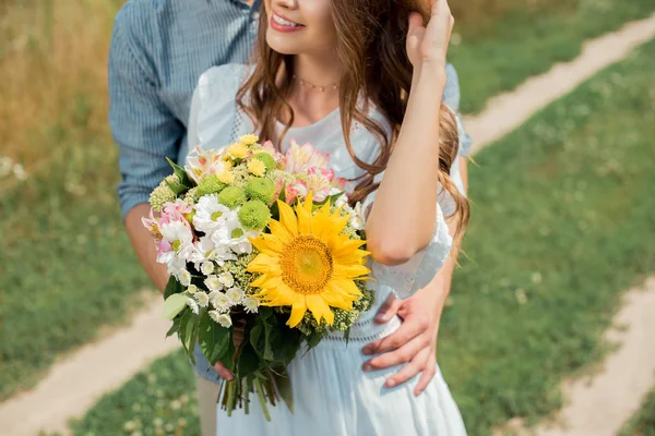 Teilansicht des Mannes umarmt lächelnde Freundin mit Strauß wilder Blumen im Sommerfeld — Stockfoto