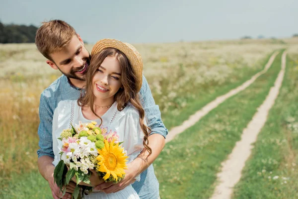 Portrait of smiling man hugging girlfriend with bouquet of wild flowers in summer field — Stock Photo