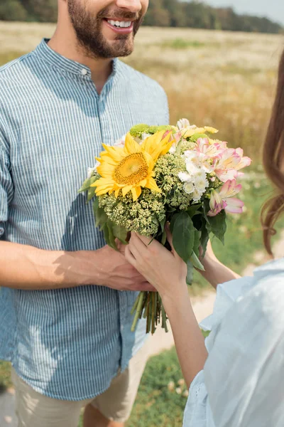 Colpo ritagliato di uomo allegro che presenta mazzo di fiori selvatici alla ragazza nel campo estivo — Foto stock