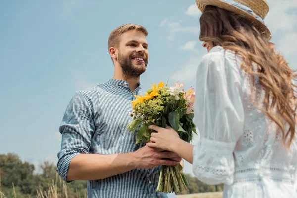 Vue à angle bas de l'homme joyeux présentant bouquet de fleurs sauvages à petite amie avec ciel bleu sur fond — Photo de stock