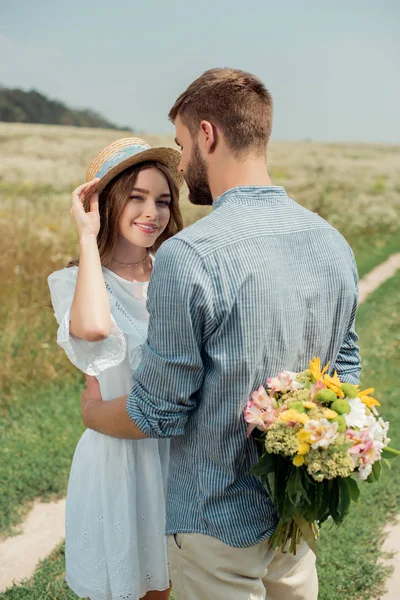 Homme cachant bouquet de fleurs sauvages pour petite amie derrière le dos dans le champ d'été — Photo de stock