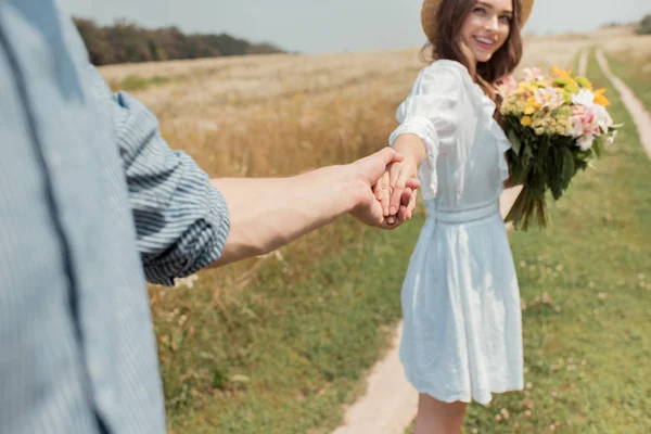 Vue partielle de femme souriante avec bouquet de fleurs sauvages et petit ami tenant la main dans le champ — Photo de stock