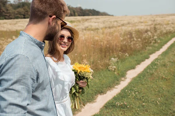 Vista lateral de mujer sonriente con ramo de flores silvestres y novio cerca en el campo - foto de stock