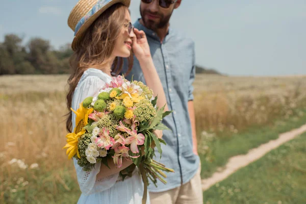 Vista lateral de mujer sonriente con ramo de flores silvestres y novio cerca en el campo - foto de stock