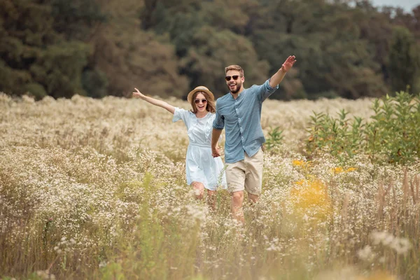 Amantes felices tomados de la mano mientras corren en el campo con flores silvestres - foto de stock