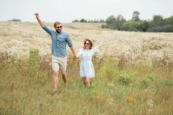 Happy lovers holding hands while running in meadow with wild flowers — Stock Photo