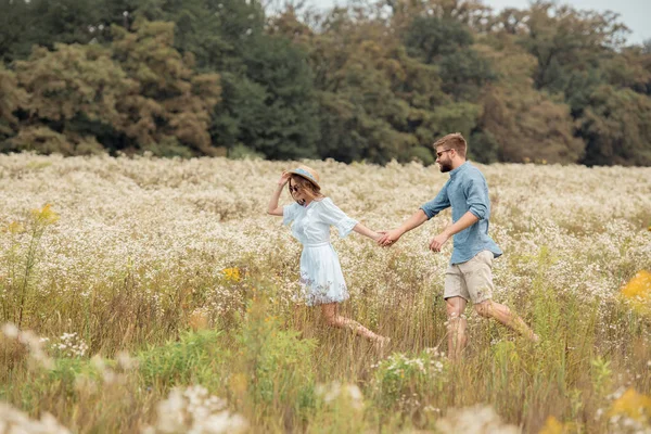Vista lateral de los jóvenes amantes tomados de la mano mientras caminan juntos en el campo con flores silvestres - foto de stock