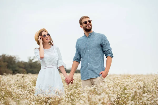 Portrait of happy lovers in sunglasses holding hands in field with wild flowers — Stock Photo