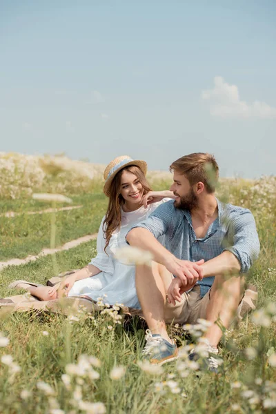 Cheerful young lovers sitting on blanket in field with wild flowers on summer day — Stock Photo