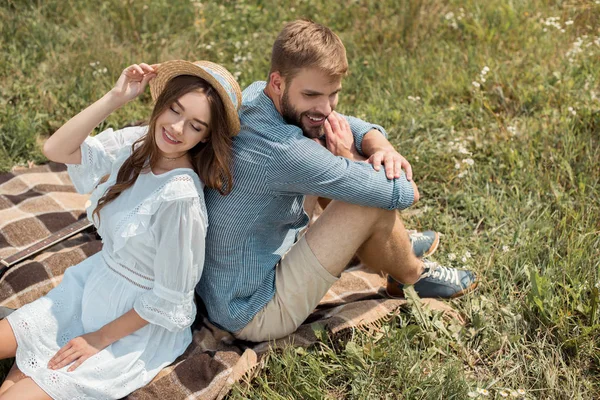 Vista de ángulo alto de la pareja sonriente descansando espalda con espalda en la manta en el campo con flores silvestres en el día de verano - foto de stock