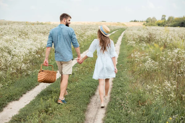 Rückansicht eines Paares mit Picknickkorb, das Händchen hält, während es im Feld geht — Stockfoto