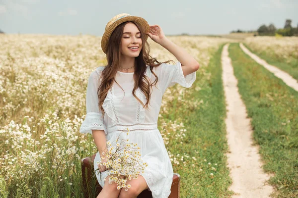 Donna sorridente in abito bianco con bouquet di fiori di camomilla selvatica seduta su valigia retrò in campo — Foto stock