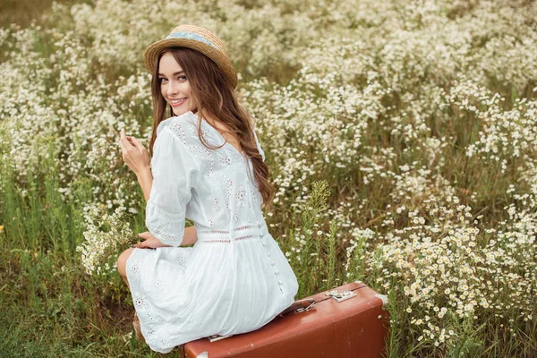 Back view of smiling woman in white dress with bouquet of wild camomile flowers sitting on retro suitcase on meadow — Stock Photo
