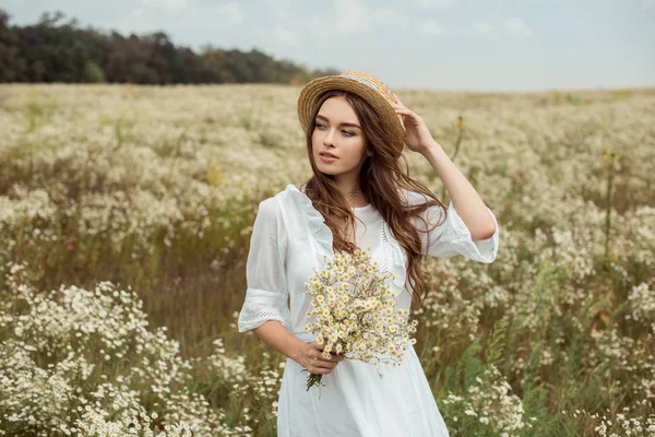 Portrait of pretty pensive woman in white dress with bouquet of wild camomile flowers on meadow — Stock Photo